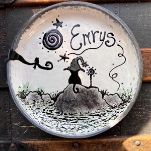 May include: A round ceramic plate with a cream-colored surface featuring a black hand-painted illustration of a cat wearing a pointed hat, a spiral moon, stars, a bird on a branch, and the name Emrys written in black script lettering.