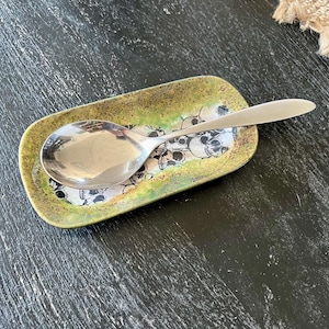 May include: A stainless steel spoon rests on a rectangular spoon rest. The spoon rest has a green and gold border with a black and white skull pattern in the center. The spoon is reflective.