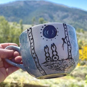 May include: A handmade ceramic mug with a blue and white speckled glaze. The mug features a black illustration of a sun, trees, and two figures. The handle is a matching blue. The background is blurred, showing a mountain and greenery.