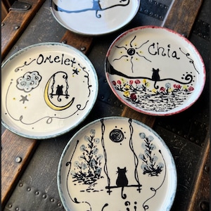May include: Four round ceramic plates featuring hand-painted black cat illustrations and names. One plate shows a cat on a line with the name Luna, another shows a cat on a swing with the name Omelet, one shows a cat with the name Chia, and one shows a cat on a swing with the name Goji.