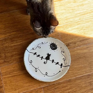 May include: A round white ceramic plate featuring a black hand-painted illustration of a cat sitting on a wire among several small birds, with a swirling sun design above. The plate has a speckled texture and a slightly uneven, rustic edge.