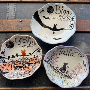 May include: Three hand-painted ceramic bowls with irregular edges. Each bowl features a unique Halloween-themed design, including cats, bats, pumpkins, and the names "Biggs", "Goblin", and "Talia". The bowls have a cream-colored base with black, orange, and purple accents.