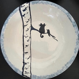 May include: A round ceramic bowl with a cream-colored interior and a blue-toned rim. The bowl features a hand-painted black illustration of a tall tree trunk with a branch extending to the right, where a cat and a small bird are perched.
