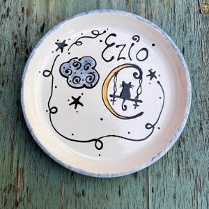 May include: A round ceramic plate with a cream-colored surface featuring a hand-painted black cat sitting on a yellow crescent moon swing. The design includes a blue cloud, black stars, and the name Ezio written in black script, all framed by a thin black line.