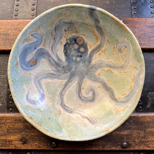 May include: A round ceramic bowl featuring a hand-painted octopus design in shades of blue and grey. The background of the bowl has a mottled, light green and tan glaze finish. The octopus is centered with swirling tentacles extending outward.