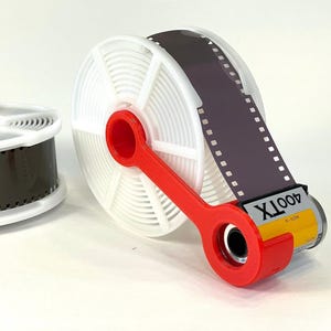 May include: A white plastic film developing reel holding a strip of dark 35mm film, connected by a bright red plastic crank arm to a yellow and silver 400TX film canister. A second white film reel with dark film sits to the left of the main assembly.