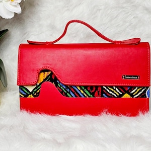 Peut inclure: Pochette en cuir rouge uni avec un accent en tissu à motifs colorés. La pochette a une poignée et une fermeture à rabat. Le texte "Ankara Touch" est visible.