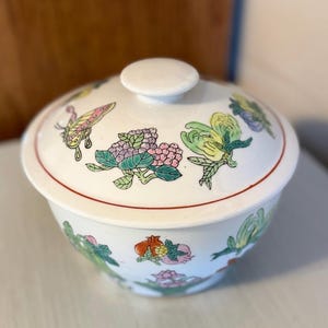 May include: White porcelain bowl with lid, featuring a floral and butterfly design in shades of green, pink, yellow, and blue. The bowl has a red rim.