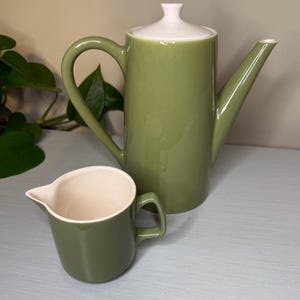Może przedstawiać: Zielony ceramiczny dzbanek do kawy z białą pokrywką i pasującym kremem. Dzbanek do kawy ma długi dzióbek i zakrzywiony uchwyt. Kremówka ma mały uchwyt i dzióbek do nalewania. Zestaw jest w jednolitym zielonym kolorze.