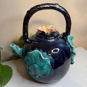 Puede incluir: Tetera de cerámica artesanal con un esmalte azul oscuro. La tetera presenta un asa marrón oscuro, una flor en la tapa y detalles florales turquesa. El pico y el asa están intrincadamente diseñados, lo que se suma a su estética única.