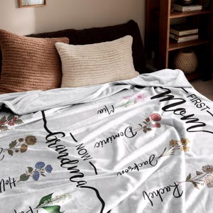 May include: White fleece blanket with floral designs and the text "First - Mom - Now - Grandma" printed on it.
