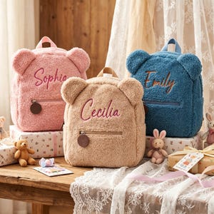 May include: Three plush bear-shaped backpacks in pink, tan, and blue. Each backpack has a zipper pocket, a top handle, and embroidered names: Sophia, Cecilia, and Emily. The backpacks are displayed on decorative boxes.