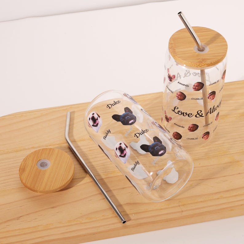 Puede incluir: Dos vasos de vidrio transparente con tapas de bamb&uacute; y pajitas de acero inoxidable. Un vaso presenta un patr&oacute;n repetido de caras de perros y los nombres "Duke" y "Buddy". El otro vaso tiene las palabras "Love & Always" y caras.