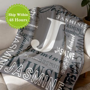 Può includere: Una coperta grigia con il nome "Jasmine" stampato in vari font e dimensioni. Una grande lettera "J" bianca è centrata sulla coperta. La coperta è drappeggiata su un divano bianco.