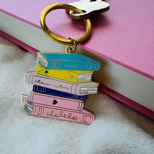 以下が含まれることがあります： カラフルなエナメル製のブックチャームが5つ重なったゴールドのキーホルダー。本はそれぞれ色が異なり、デザインも異なります。青い本には波線、黄色の本には星、ピンクの本にはハート、青い本には「本が好き」という文字が書かれています。