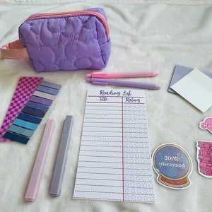 Puede incluir: Un estuche de lápices acolchado morado con un asa rosa, una lista de lectura con una columna de título y calificación, dos resaltadores rosa y morado, un conjunto de notas adhesivas de colores y varias pegatinas con temática de libros. Las pegatinas incluyen un corazón con el texto "Leer más libros", una pila de libros con el texto "Nunca puedes tener demasiados libros", y una bola de cristal con el texto "Obsesionado con los libros".