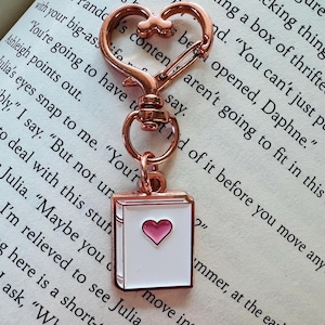 Puede incluir: Un llavero de oro rosa con un cierre en forma de corazón y un dije de esmalte blanco con forma de libro con un corazón rosa en la portada. El dije está unido a una cadena de oro rosa.