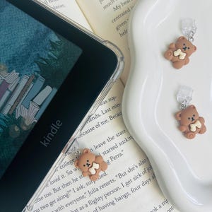 Könnte beinhalten: Zwei braune Handy-Anhänger in Bärenform mit einem weißen Knochen-Accessoire. Die Anhänger sind an durchsichtigen Plastik-Handygriffen befestigt.