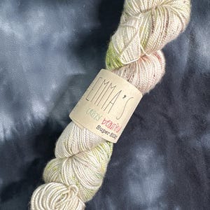 May include: A skein of Emma's Crazy Beautiful Super Silky yarn. The yarn is a blend of cream, light pink, and green, with a speckled appearance. The label is a light brown color with the brand name and yarn type printed on it.