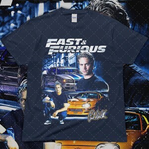 Puede incluir: Camiseta azul marino con el texto "Fast & Furious" y im&aacute;genes de coches y Paul Walker. El dise&ntilde;o incluye un coche plateado con rayas azules y un coche naranja. La camiseta tiene un cuello redondo cl&aacute;sico.
