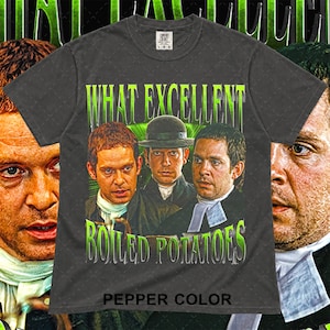 May include: A charcoal gray short-sleeve t-shirt featuring a graphic print of three men in period attire. The text "WHAT EXCELLENT BOILED POTATOES" is printed in bright green lettering above and below the figures. The shirt has a crew neckline.