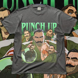 May include: Dark gray t-shirt featuring a graphic of a man with a large mustache and the text "PUNCH UP" in green. The graphic includes multiple images of the man in various poses, some with tattoos. The shirt has a vintage, worn look.