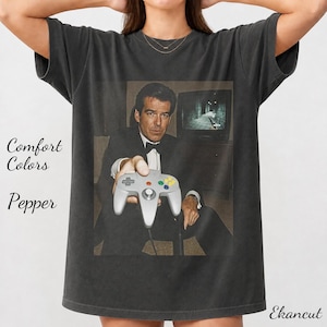May include: A charcoal grey t-shirt featuring a graphic print of a man in a black tuxedo holding a grey video game controller. The print includes a television screen in the background. The text Comfort Colors Pepper and Ekancut is visible.