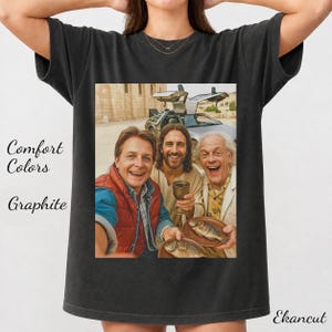 May include: A graphite-colored t-shirt featuring a graphic print of three individuals, including one holding a chalice and another holding a plate with fish, with a silver sports car with gull-wing doors in the background. The text Comfort Colors and Graphite is visible.