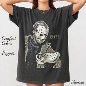 Könnte beinhalten: Ein anthrazitfarbenes Kurzarm-T-Shirt mit einem Grafikdruck einer Figur mit Hockeymaske, die einen weißen, perforierten Clog hält. Der Text EH?? ist in einer hellgrauen Schriftart neben der Figur aufgedruckt. Das Shirt hat einen Rundhalsausschnitt.