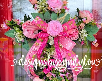Op de afbeelding: Een levendige bloemenkrans met grote roze rozen, kleinere roze bloemen en groen blad. De krans is versierd met roze linten, waaronder stippen- en bloempatronen. De tekst "designsbycindylow" is zichtbaar.