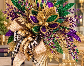 May include: A decorative centerpiece featuring a gold pedestal base, a central glittered flower in purple, green, and gold, topped with a diamond-patterned finial. It includes black and white checkered ribbon, gold patterned ribbon, and foliage.