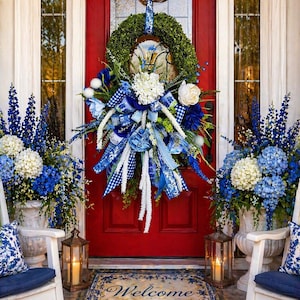 May include: A vibrant red front door adorned with a lush green wreath and a large blue and white floral arrangement. Matching blue and white floral arrangements flank the door, with a welcome mat and lanterns completing the welcoming scene.