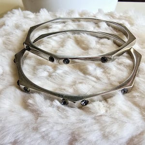 May include: Two silver bangle bracelets with black rhinestone accents. The bracelets are stacked on top of each other.