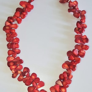 Könnte beinhalten: Eine leuchtend rote Halskette aus unregelmäßig geformten Korallenstücken. Die Halskette hat eine kleine Perlenkette und einen Verschluss. Die Korallenstücke sind gruppiert, was ein auffälliges Accessoire ergibt.