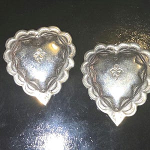 May include: Two silver-toned heart-shaped conchos with a scalloped edge and a central floral design. The conchos have a textured surface and a pointed bottom. They are likely used for decorative purposes on leather goods or clothing.