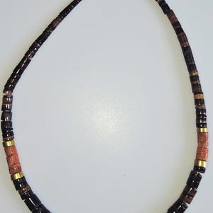 May include: A beaded necklace featuring a variety of dark brown, black, and reddish-brown beads. The necklace has a silver clasp and includes gold-colored spacers. The beads are rectangular and cylindrical.