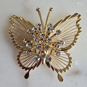 May include: A gold butterfly brooch with clear rhinestones. The butterfly has a wire frame design and is decorated with a cluster of rhinestones in the center.