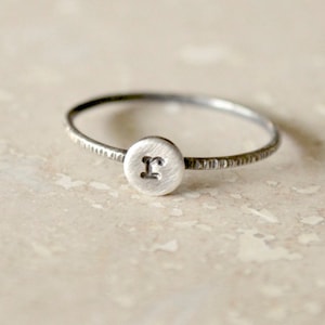 May include: A silver ring with a textured band and a round disc featuring the letter "F" engraved on it.