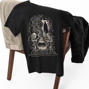 Op de afbeelding: Een zwart t-shirt met een witte grafische illustratie van drie figuren rond een ketel en een silhouet op de achtergrond. De tekst luidt: BY THE PRICKING OF MY THUMBS, SOMETHING WICKED THIS WAY COMES. WILLIAM SHAKESPEARE - MACBETH - ACT IV.
