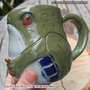 Pode incluir: Uma caneca de cerâmica esculpida no formato de uma rã-de-olhos-vermelhos verde, com um olho vermelho proeminente, barriga branca e uma seção com padrão azul na lateral. O texto ART BY FENIX | MANICGREMLIN.COM e SCULPTURAL RED EYED TREE FROG MUG é visível.