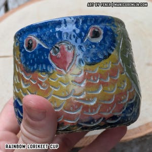 May include: A ceramic cup featuring a hand-painted rainbow lorikeet design with a blue face, red beak, and yellow, orange, and blue feathers. The surface has carved texture details. Text on the image reads ART BY FENIX | MANICGREMLIN.COM and RAINBOW LORIKEET CUP.