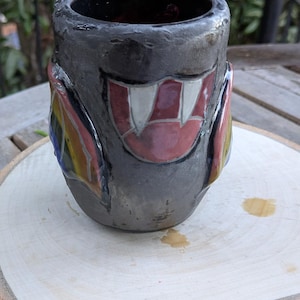 May include: A handmade ceramic mug with a dark gray exterior and a colorful, stylized design. The mug features a red, white, and pink mouth design with white fangs. The text "Pride Potsferatu #1" is at the bottom. The mug is on a wooden surface.