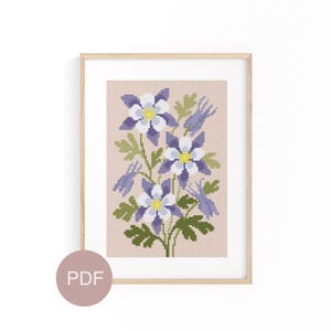 May include: A framed cross-stitch floral design featuring blue and white flowers with yellow centers, green leaves, and blue buds. The artwork is set against a light pink background and framed in a light wood frame. A PDF icon is in the lower left corner.