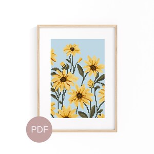 May include: A cross-stitch pattern featuring a bouquet of yellow flowers with brown centers on a light blue background. The pattern is framed in a light wood frame.
