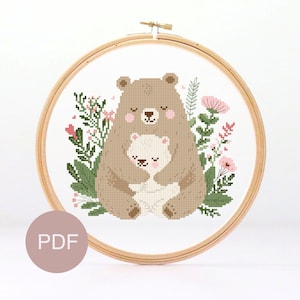 May include: Cross stitch pattern featuring a brown bear hugging a white bear cub, surrounded by green leaves and pink flowers. The pattern is in a circular embroidery hoop.
