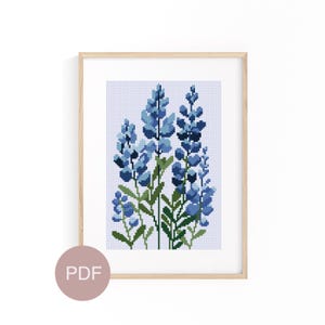 May include: A framed cross-stitch of bluebonnet flowers. The artwork features a light blue background with a grid pattern. The flowers are various shades of blue with green stems and leaves. The frame is light wood.