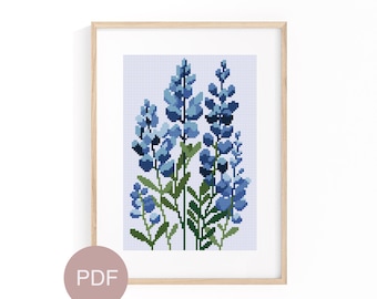 May include: A framed cross-stitch of bluebonnet flowers. The artwork features a light blue background with a grid pattern. The flowers are various shades of blue with green stems and leaves. The frame is light wood.