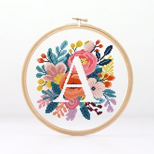 May include: Cross-stitch embroidery of the letter "A" surrounded by colorful flowers and foliage. The design features pink, orange, yellow, and teal elements. The embroidery is set in a wooden hoop, creating a decorative piece.