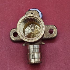 May include: A brass plumbing fitting with a threaded interior and a series of stacked cylindrical sections. The fitting has two side extensions with holes and a silver-colored top piece. The background is a solid red color.