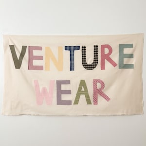 May include: A rectangular off-white fabric banner featuring the text VENTURE WEAR appliqued in various patterned and solid-colored fabric letters. The letters include green, pink, yellow, blue, black, and white plaid, gingham, and floral prints.